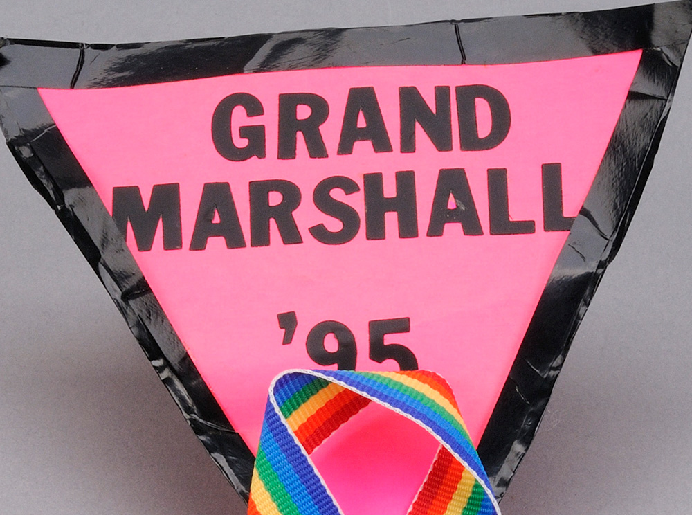 Zoom d'une épinglette triangulaire en tissu rose avec un ruban arc-en-ciel en bas et les mots « Grand Marshall, “95 » Close-up of a pink fabric triangle pin with a rainbow ribbon at the bottom, and the words “Grand Marshall, ’95.”
