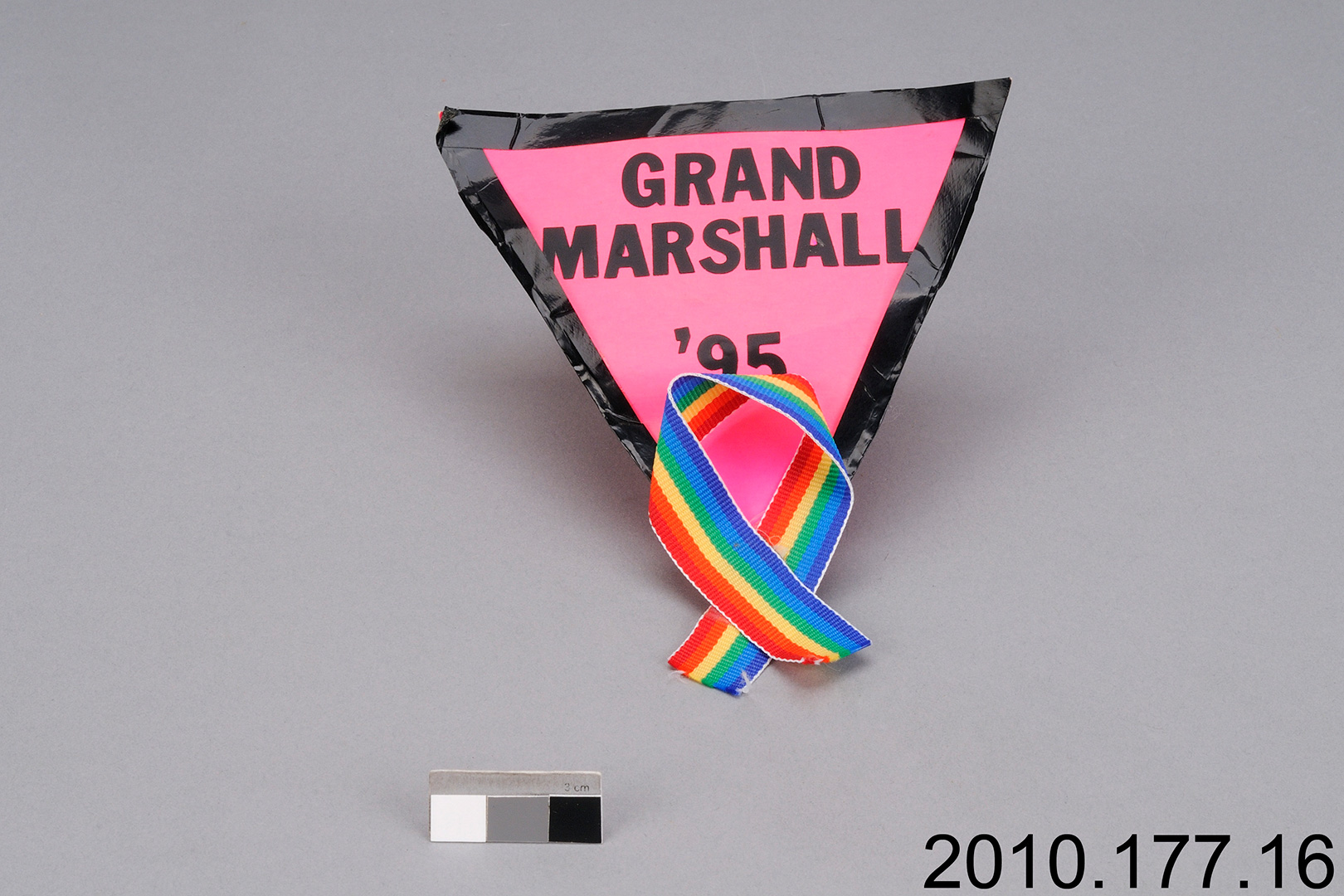 Une épinglette triangulaire en tissu rose avec un ruban arc-en-ciel en bas et les mots « Grand Marshall, “95 » A pink fabric triangle pin with a rainbow ribbon at the bottom, and the words “Grand Marshall, ’95.”