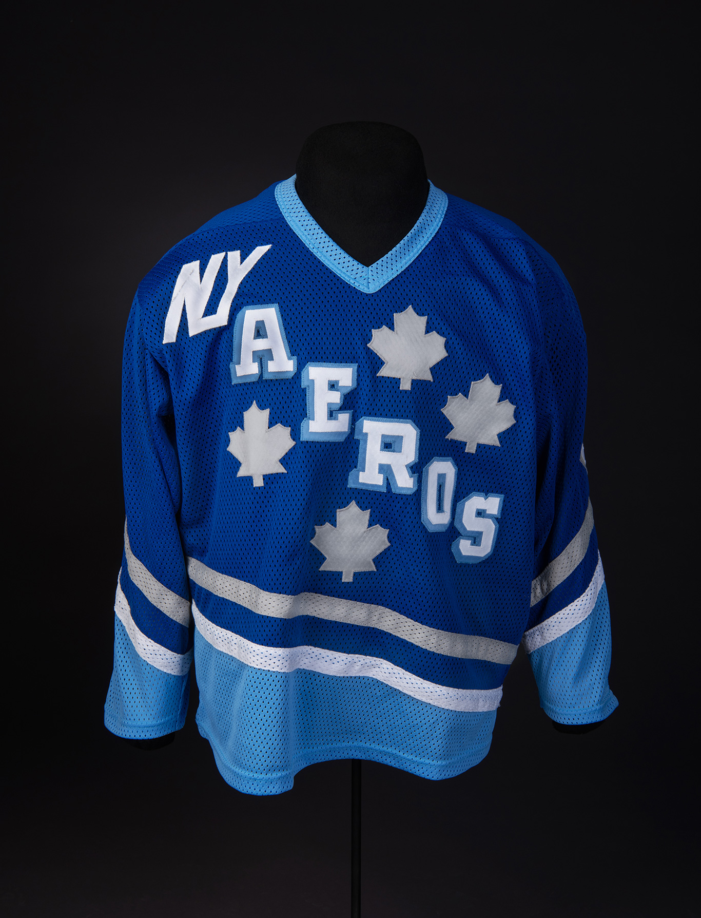 : Maillot de hockey bleu avec l’inscription NY AEROS en lettres blanches et 4 feuilles d’érable blanches autour des lettres. Blue hockey jersey reads “NY AEROS” in white lettering with four white maple leaves around the lettering.