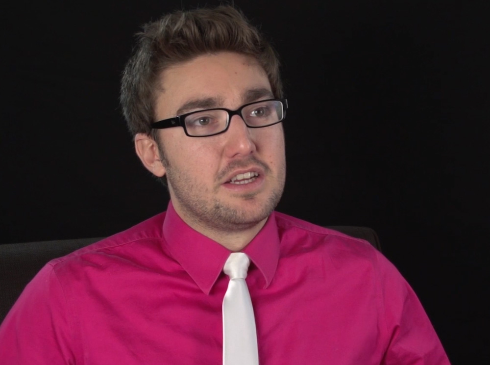 A screenshot of a video clip featuring Travis Price, a man with glasses wearing a pink shirt and white tie.
