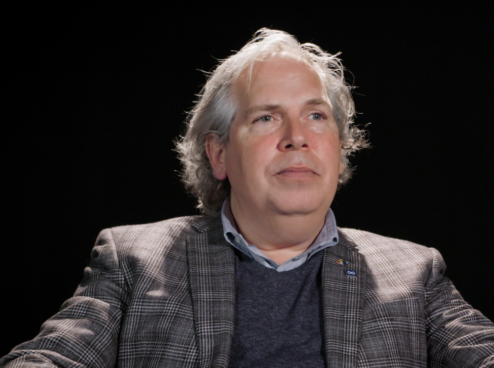 A screenshot of a video clip featuring Todd Ross, seated wearing a brown suit.