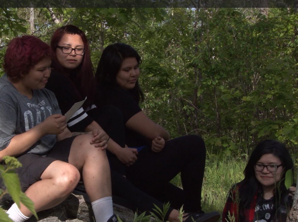 Four Indigenous teenagers sitting on a rock in a forest.