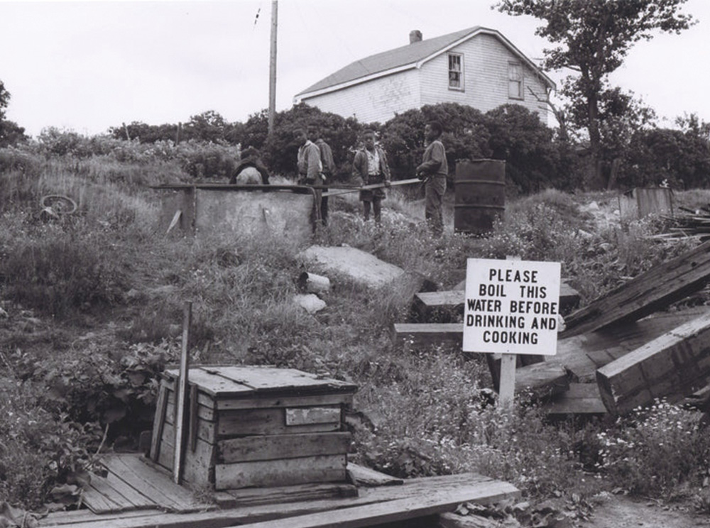 Black-and-white photograph of a group of children playing outside near a railway car, and a large sign with a boil-water advisory in the foreground.