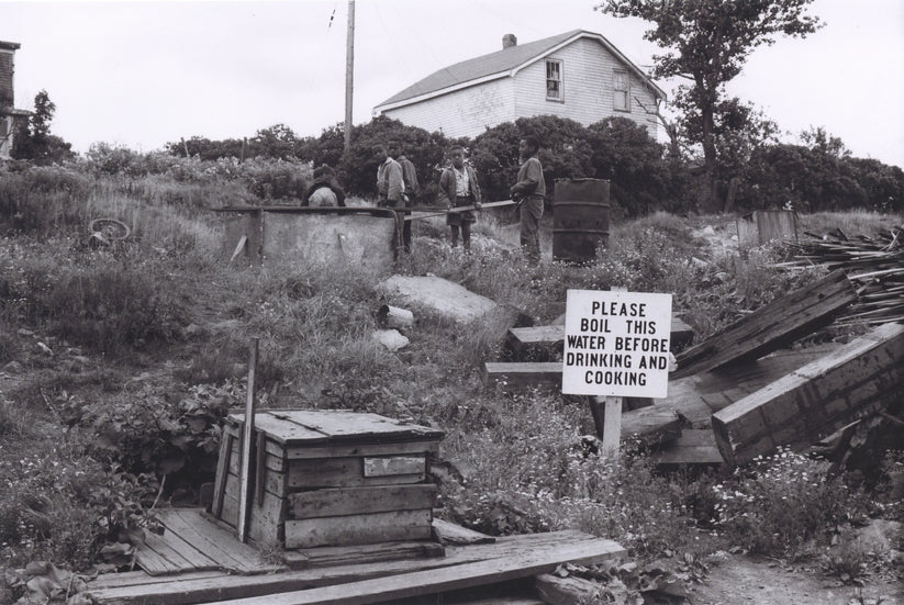Photographie en noir et blanc d’un groupe d’enfants jouant à l’extérieur près d’un wagon de chemin de fer, et d’un grand panneau avec un avis d’ébullition de l’eau au premier plan.
