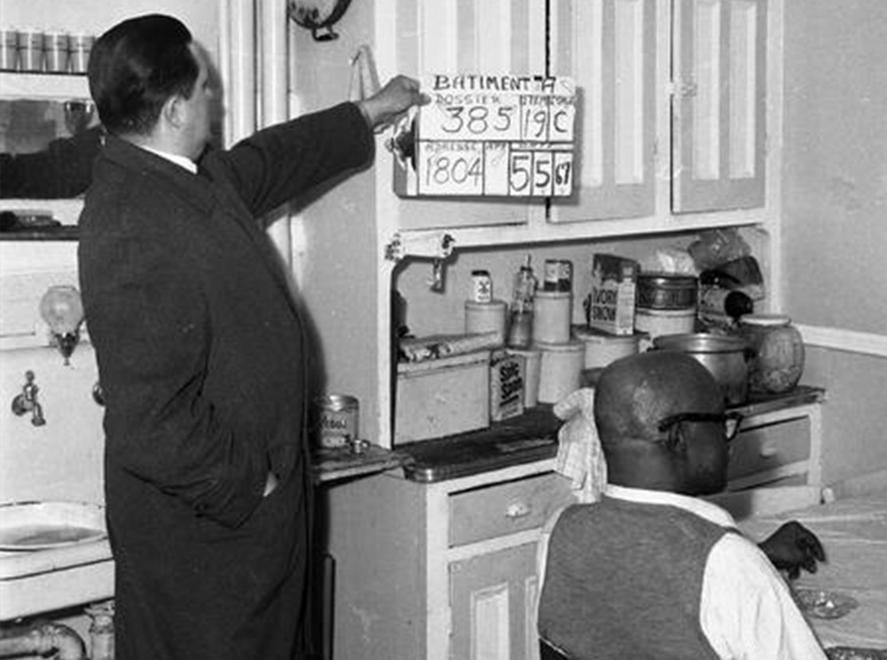 Black and white photograph of a city official in the kitchen of a home with a man seated at the kitchen table