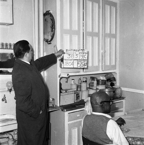 Photographie en noir et blanc d’un fonctionnaire municipal dans la cuisine d’une maison avec un homme assis à la table de la cuisine