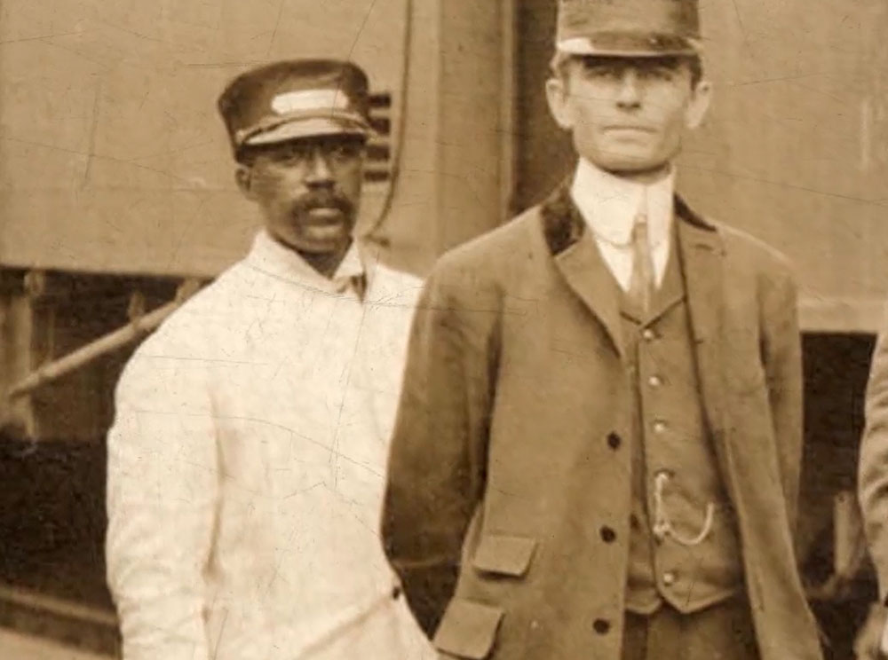 Screen capture from a video of a white man in a tan uniform standing in front of a black man in a white uniform.