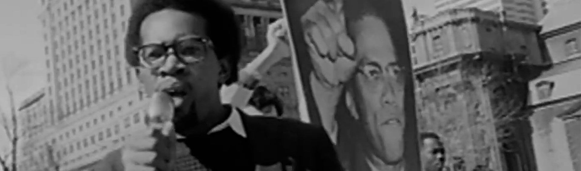 Black-and-white photograph of a Black man speaking into a microphone during a protest march.