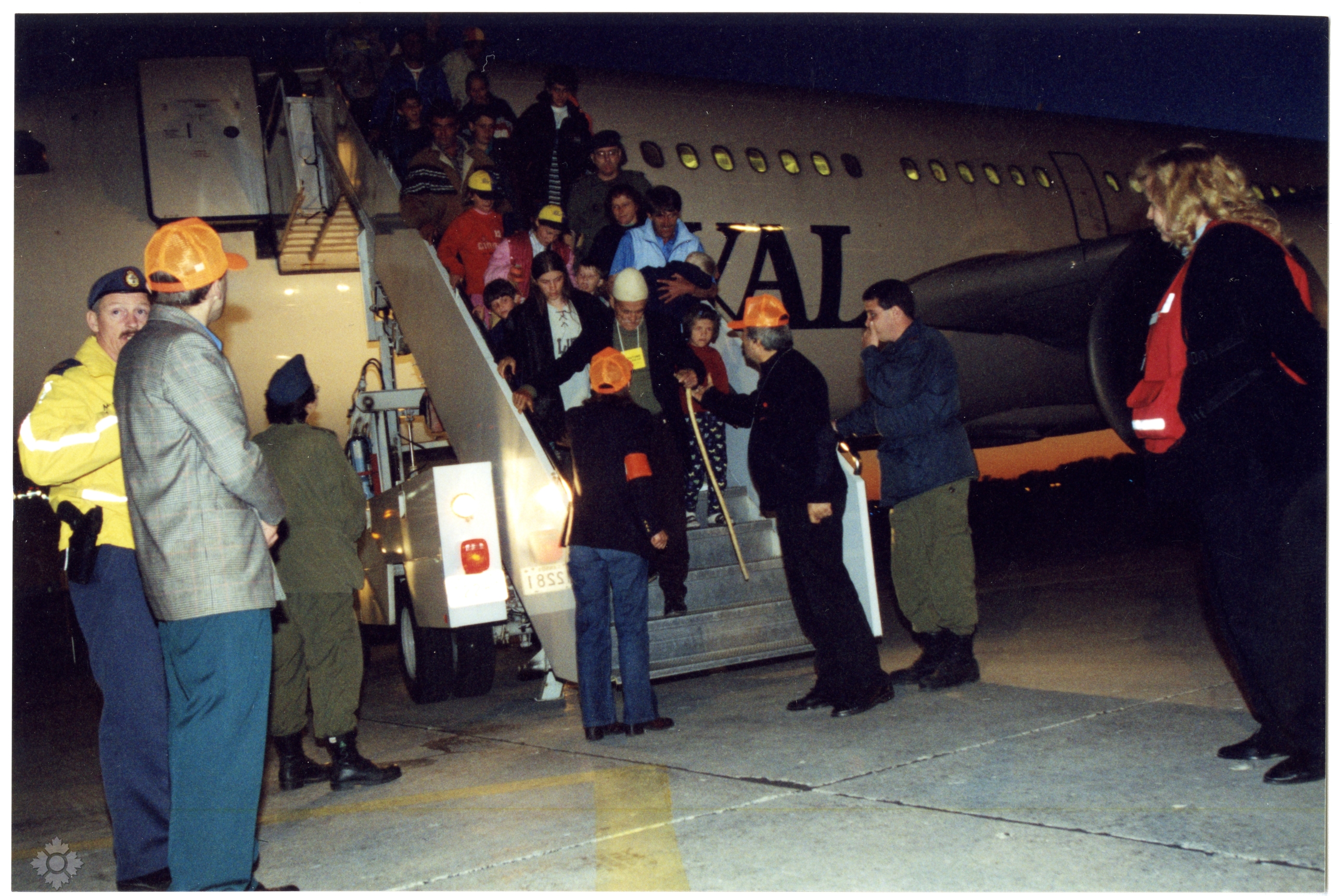 Des personnes réfugiées kosovares descendent d’un avion en Ontario en 1999. Elles sont accueillies par des interprètes portant des casquettes orange, des membres de la Croix-Rouge et des militaires.