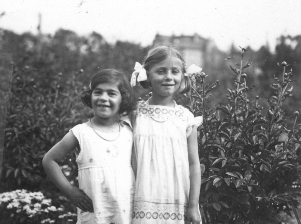 Black-and-white photograph of two light-skinned young girls in short white dresses, smiling and standing close together in a garden, next to a hedge.