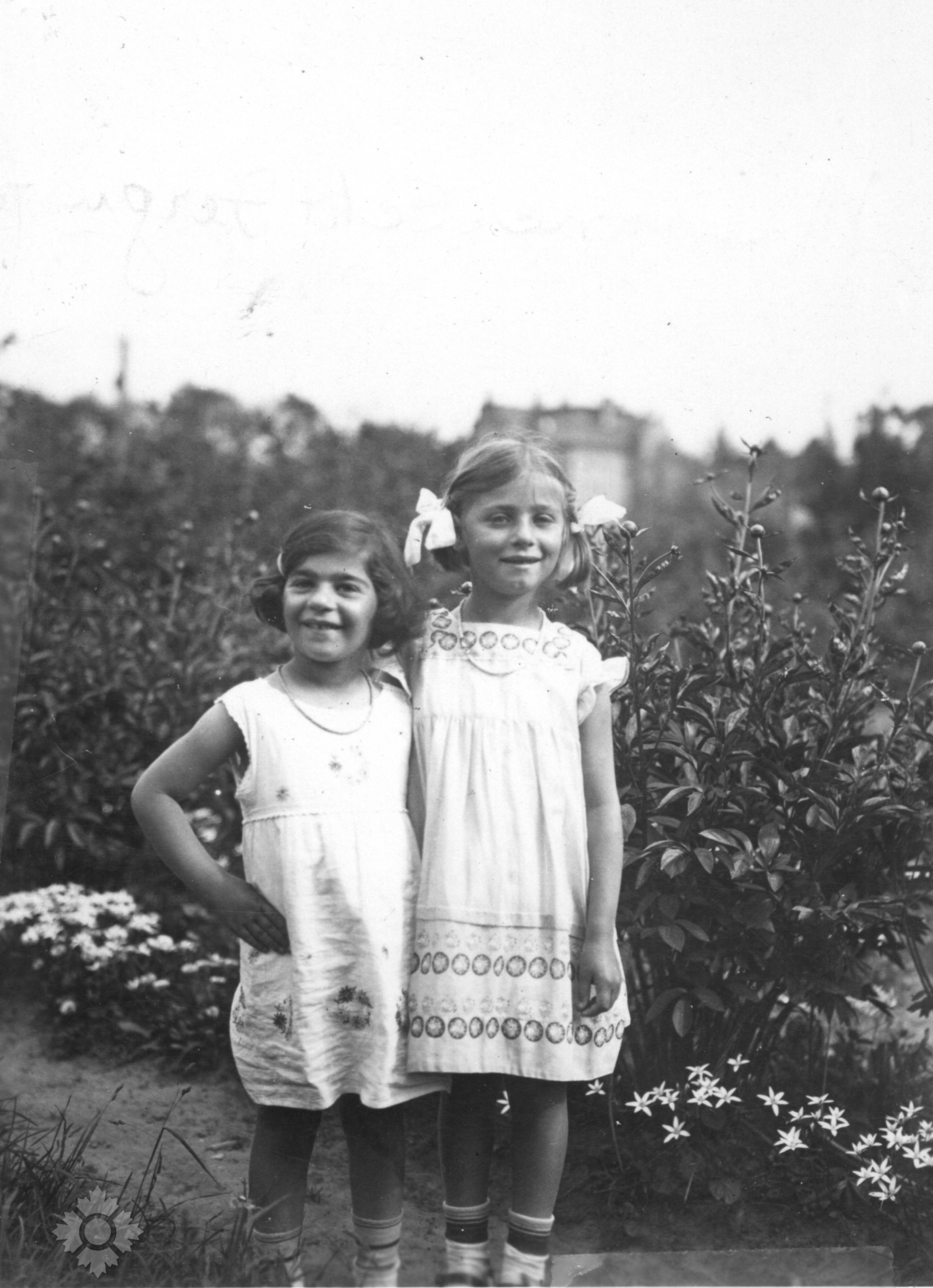 Black-and-white photograph of two light-skinned young girls in short white dresses, smiling and standing close together in a garden, next to a hedge.
