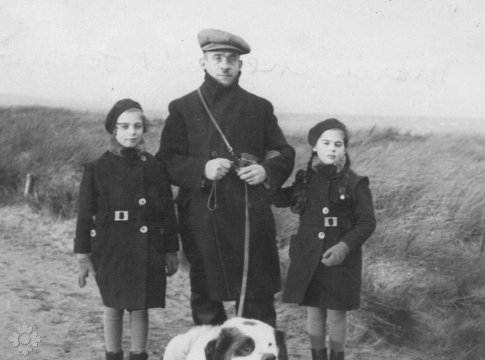 Une photographie en noir et blanc d’un homme à la peau claire tenant un chien blanc en laisse, avec, à ses côtés, deux filles à la peau claire. Ils sont dehors et portent tous un manteau d’hiver d’une couleur foncée.