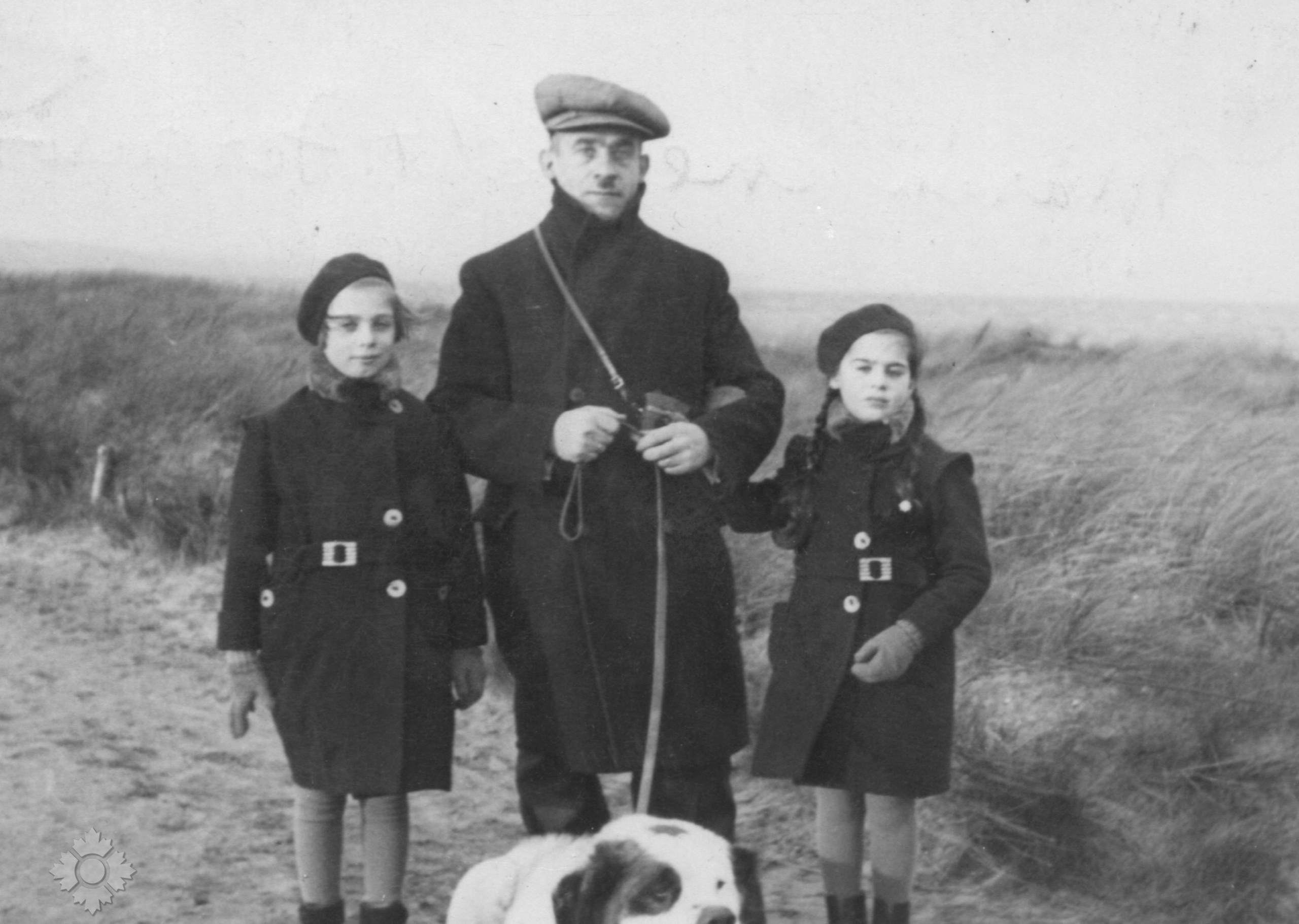 Une photographie en noir et blanc d’un homme à la peau claire tenant un chien blanc en laisse, avec, à ses côtés, deux filles à la peau claire. Ils sont dehors et portent tous un manteau d’hiver d’une couleur foncée.