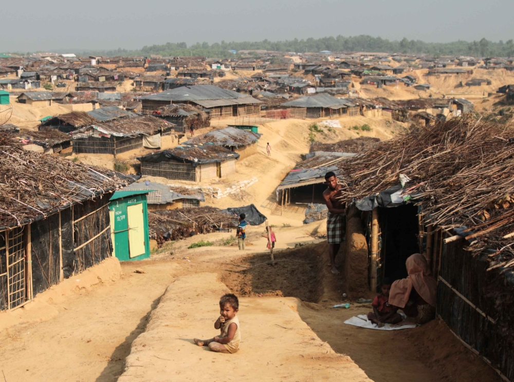 Colour photograph of a small dark-skinned child sitting on a dirt path between huts made of branches and grasses. There are many more huts in the background.