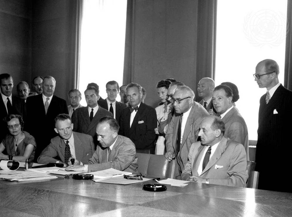 A black-and-white photograph of about 20 men in suits. Some are sitting at a table signing documents. Others stand behind them, looking on.