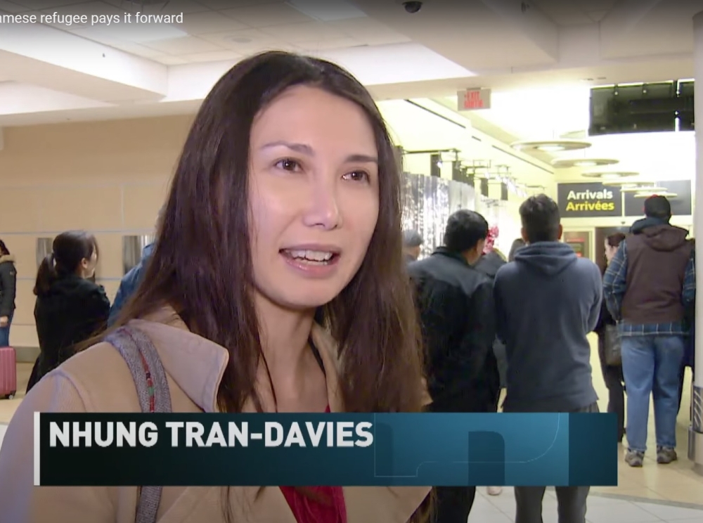 A Vietnamese woman with long dark hair and a beige coat, smiling and talking to someone off-camera in a large indoor area with people walking around in the background.