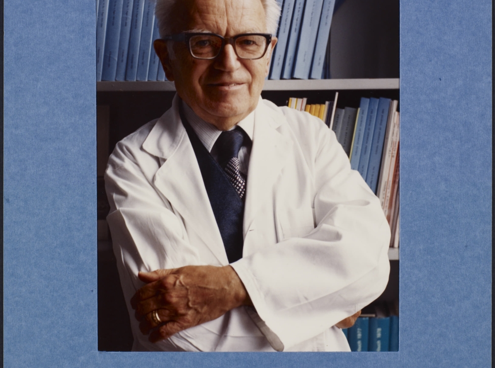Une photographie en couleur d’un homme souriant d’un certain âge à la peau claire et aux cheveux blancs qui porte des lunettes et une blouse blanche, et a les bras croisés.
