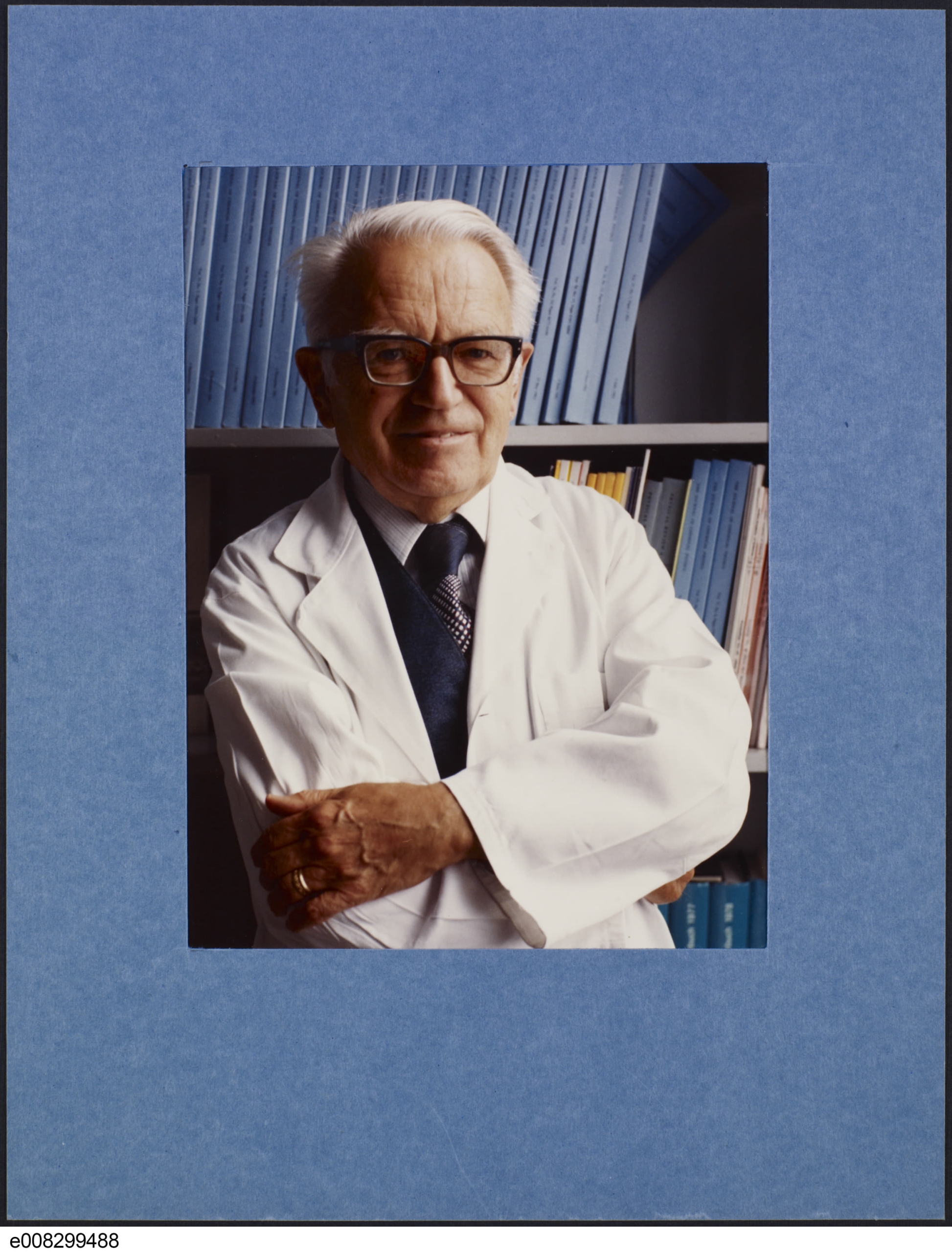 Une photographie en couleur d’un homme souriant d’un certain âge à la peau claire et aux cheveux blancs qui porte des lunettes et une blouse blanche, et a les bras croisés.