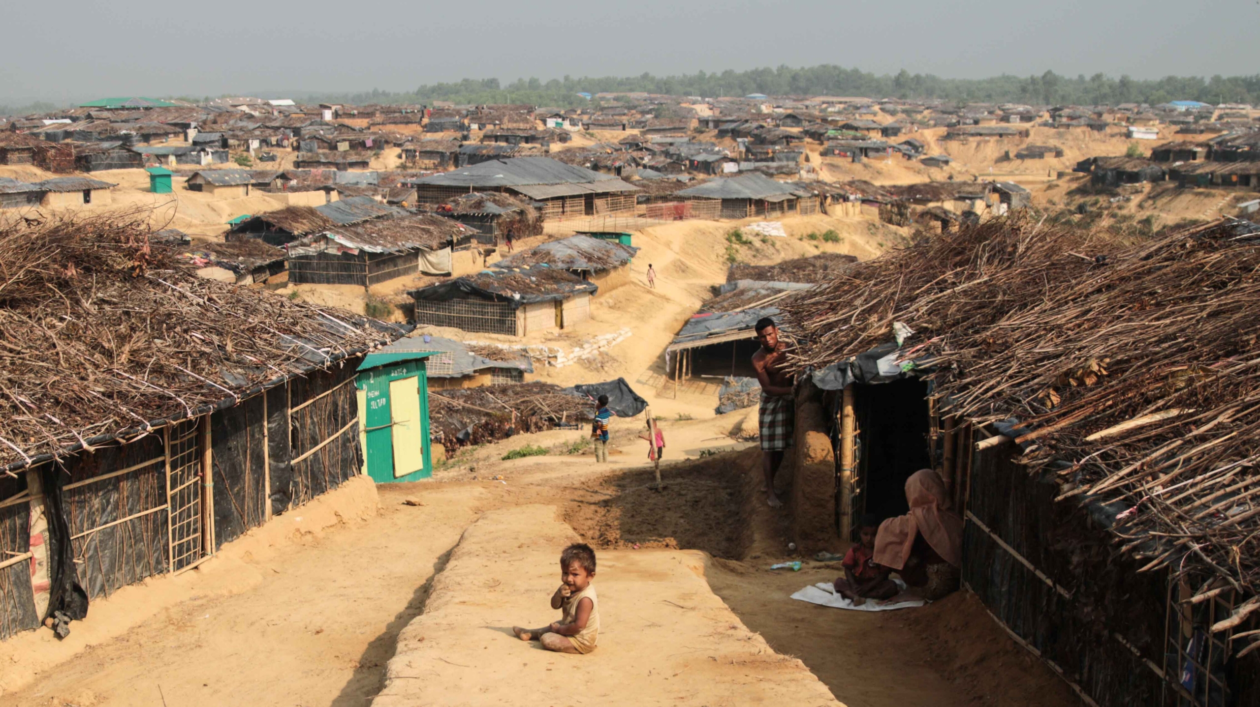 Une photographie en couleur d’un petit enfant à la peau foncée assis par terre entre des huttes construites avec des branches et du chaume, et séparées par un chemin de terre. Il y a de nombreuses huttes à l’arrière-plan. 