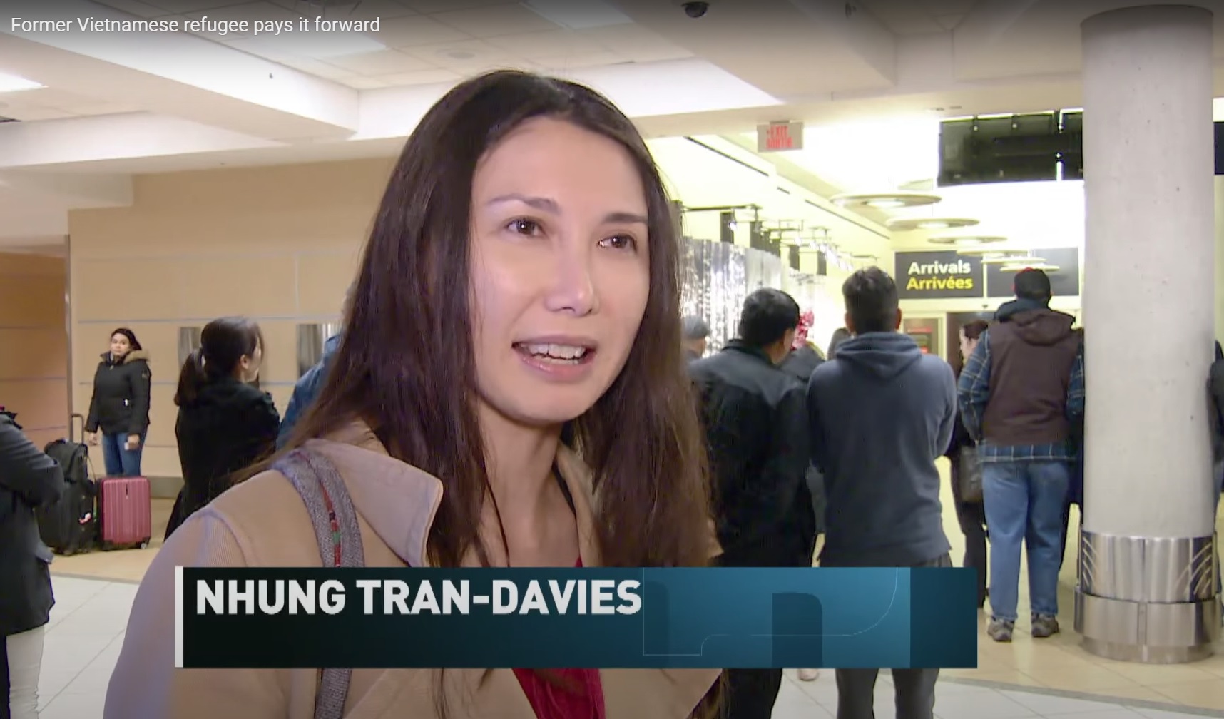 Une Vietnamienne aux cheveux bruns longs portant un manteau beige, souriant et parlant à quelqu’un qui est derrière la caméra dans un grand espace intérieur avec des personnes qui marchent à l’arrière-plan.