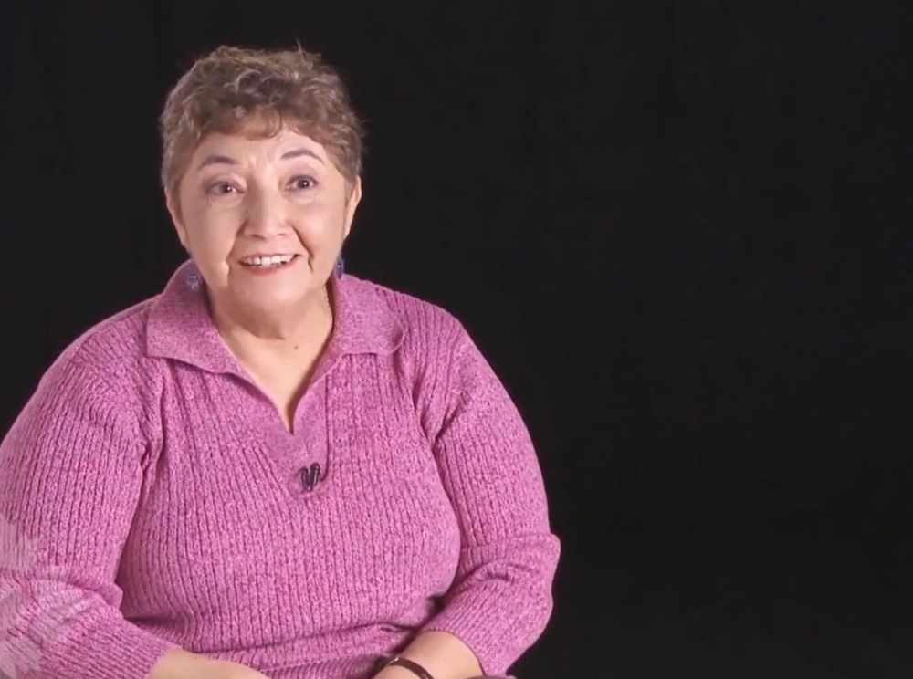 Une image fixe tirée d’une vidéo montrant une femme au teint moyen et aux cheveux courts qui porte un haut rose foncé et sourit à la caméra sur un fond sombre.